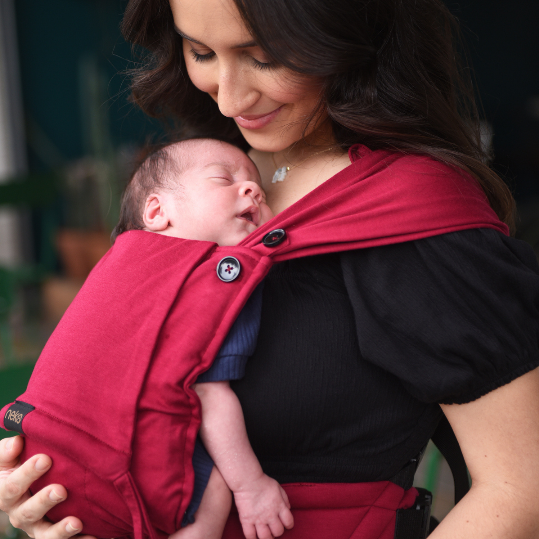Eltern tragen ihr Neugeborenes in der Neko Tiny Babytrage in Sangria – sanft, ergonomisch und anschmiegsam.